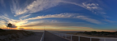 A panoramic view of a boardwalk leading to an expansive beach, with a wooden hut on stilts to the left and a vast sky painted with dramatic, sweeping clouds during sunset.