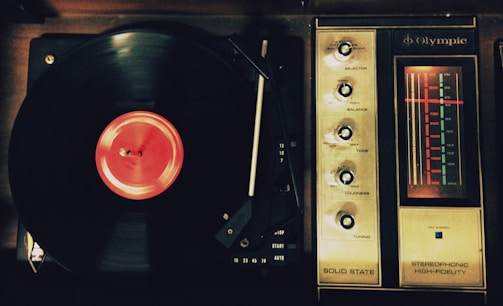 A vintage record player with a spinning vinyl record on the left side. The record has a bright red label. To the right, an analog radio tuner with dials for selector, balance, tone, loudness, and tuning is visible. The tuner displays frequencies with an illuminated red indicator.