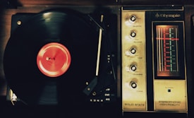 A vintage record player with a spinning vinyl record on the left side. The record has a bright red label. To the right, an analog radio tuner with dials for selector, balance, tone, loudness, and tuning is visible. The tuner displays frequencies with an illuminated red indicator.