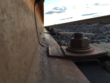A close-up view of a railway track, focusing on the metallic rail and a large bolt securing it. The perspective captures the rail extending into the distance, with gravel stones scattered alongside. The sky above is slightly cloudy, indicating an evening or late afternoon setting.