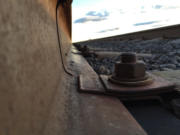 A close-up view of a railway track, focusing on the metallic rail and a large bolt securing it. The perspective captures the rail extending into the distance, with gravel stones scattered alongside. The sky above is slightly cloudy, indicating an evening or late afternoon setting.