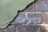 A section of crumbling plaster wall partially revealing the older brickwork underneath. The plaster is painted green and is peeling away to show dusty, weathered bricks with remnants of the previous white paint.