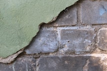 A section of crumbling plaster wall partially revealing the older brickwork underneath. The plaster is painted green and is peeling away to show dusty, weathered bricks with remnants of the previous white paint.