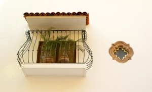 An ornate balcony with black wrought iron railing adorned with potted green plants. The balcony has a small roof with red clay tiles. Adjacent to the balcony is a decorative round window with an intricate border carved in stone. The wall is painted light yellow or off-white.