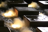 Large stone blocks are set in a water feature with illuminated water flowing around them, creating a dynamic and vibrant display. The lighting casts a warm glow on the water, adding a sense of movement and energy.