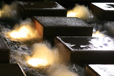 Large stone blocks are set in a water feature with illuminated water flowing around them, creating a dynamic and vibrant display. The lighting casts a warm glow on the water, adding a sense of movement and energy.