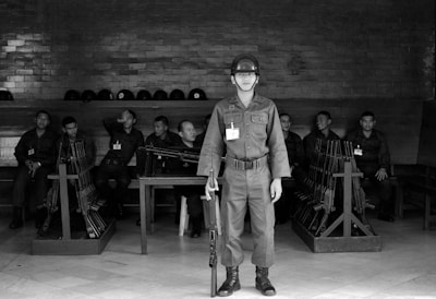 A group of soldiers is seated on benches in a dimly lit room. Each soldier is wearing a uniform and name tag. Helmets are placed on a shelf above their heads. Several racks of weapons are present near the soldiers. One soldier stands in the foreground, holding a rifle, and is positioned in the center of the scene.