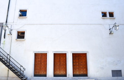 A large white building facade featuring three wooden doors at the bottom with grid patterns. Two small rectangular windows are visible at the top alongside a pair of wall-mounted lamps. An exterior staircase on the left side leads to an upper level.
