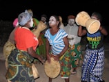 A group of people with painted faces engage in an energetic and cultural dance, holding and playing traditional percussion instruments. They are dressed in vibrant, patterned clothing, enhancing the festive atmosphere.