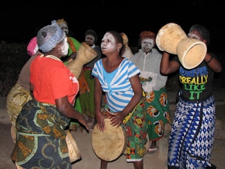 A group of people with painted faces engage in an energetic and cultural dance, holding and playing traditional percussion instruments. They are dressed in vibrant, patterned clothing, enhancing the festive atmosphere.