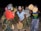 A group of people with painted faces engage in an energetic and cultural dance, holding and playing traditional percussion instruments. They are dressed in vibrant, patterned clothing, enhancing the festive atmosphere.