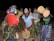 A group of people with painted faces engage in an energetic and cultural dance, holding and playing traditional percussion instruments. They are dressed in vibrant, patterned clothing, enhancing the festive atmosphere.