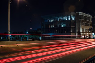 A nighttime urban scene captures a building with modern architecture, featuring large glass windows and brick exterior. Bright red and white light trails from vehicles on the road create dynamic lines across the image, indicating motion. Streetlights illuminate the area, adding to the ambient light.