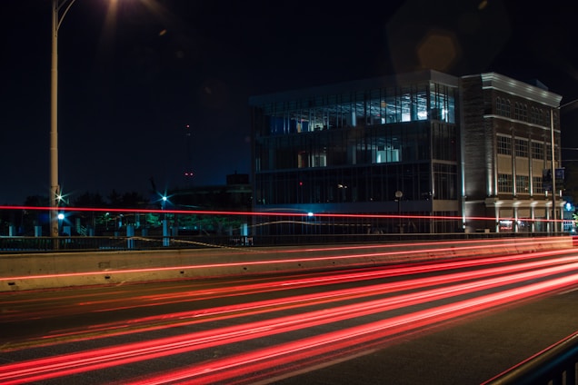 A nighttime urban scene captures a building with modern architecture, featuring large glass windows and brick exterior. Bright red and white light trails from vehicles on the road create dynamic lines across the image, indicating motion. Streetlights illuminate the area, adding to the ambient light.