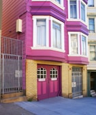 A brightly colored residential building with magenta and yellow brick exterior. The building features large windows with white frames and a double garage door painted in magenta with 'NO PARKING' signs. Metal gates and a concrete staircase lead to the entrance.