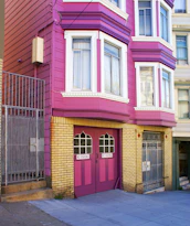 A brightly colored residential building with magenta and yellow brick exterior. The building features large windows with white frames and a double garage door painted in magenta with 'NO PARKING' signs. Metal gates and a concrete staircase lead to the entrance.