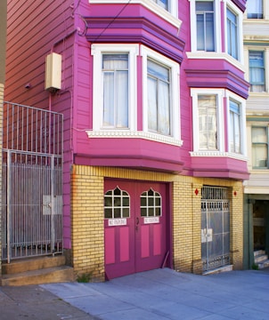 A brightly colored residential building with magenta and yellow brick exterior. The building features large windows with white frames and a double garage door painted in magenta with 'NO PARKING' signs. Metal gates and a concrete staircase lead to the entrance.