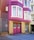 A brightly colored residential building with magenta and yellow brick exterior. The building features large windows with white frames and a double garage door painted in magenta with 'NO PARKING' signs. Metal gates and a concrete staircase lead to the entrance.