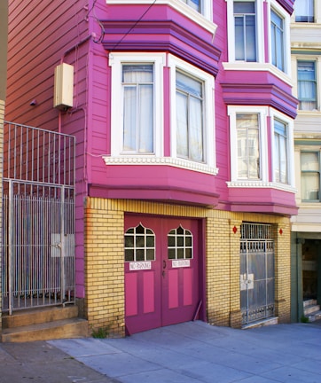 A brightly colored residential building with magenta and yellow brick exterior. The building features large windows with white frames and a double garage door painted in magenta with 'NO PARKING' signs. Metal gates and a concrete staircase lead to the entrance.