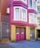 A brightly colored residential building with magenta and yellow brick exterior. The building features large windows with white frames and a double garage door painted in magenta with 'NO PARKING' signs. Metal gates and a concrete staircase lead to the entrance.