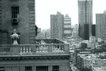 The image features a cityscape with tall buildings in the background, showcasing an urban environment. A foreground terrace with decorative railings and a large urn is visible, adding classic architectural elements to the scene.