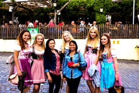 A group of seven women standing on a cobblestone street, with a wooden fence and greenery in the background. The women are smiling and wearing colorful traditional dresses, with one in casual wear. They are standing close together, seemingly enjoying a festive or cultural event.