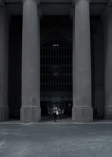 A solitary figure walks towards a building entrance framed by massive stone columns. The scene is set in an urban environment, with the grandeur of the architecture conveying a sense of history and power.
