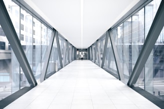 A long, modern pedestrian bridge with glass walls and a tiled walkway, featuring structural beams and city buildings visible through the windows.