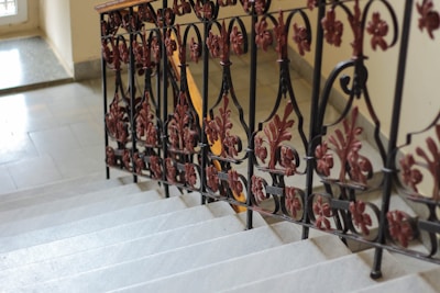An ornate metal railing with intricate designs featuring floral motifs in a dark red color lines the edge of a gray stone staircase. The steps appear worn, indicating their age, and lead downwards with a slightly glossy surface. Sunlight fills a portion of the space, highlighting the textures and details of the metalwork and the stairs.