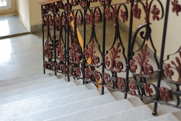An ornate metal railing with intricate designs featuring floral motifs in a dark red color lines the edge of a gray stone staircase. The steps appear worn, indicating their age, and lead downwards with a slightly glossy surface. Sunlight fills a portion of the space, highlighting the textures and details of the metalwork and the stairs.