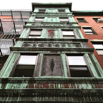 A multi-story building façade with aging green copper cladding, featuring ornate decorative designs between window frames. The structure is next to a red brick building with fire escape stairs.