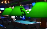 A pool table is centered in a dimly lit room with several green lampshades hanging above it. One of the lampshades closest to the camera has the number 5 on it. There are bar stools and tables in the background, suggesting the setting is a bar or pub. Background elements include wall-mounted televisions, a neon sign, and various posters or signs.