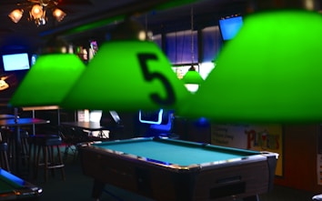 A pool table is centered in a dimly lit room with several green lampshades hanging above it. One of the lampshades closest to the camera has the number 5 on it. There are bar stools and tables in the background, suggesting the setting is a bar or pub. Background elements include wall-mounted televisions, a neon sign, and various posters or signs.
