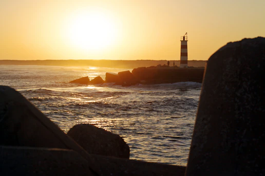 A calm ocean scene at dawn with a lighthouse casting golden light over the water.