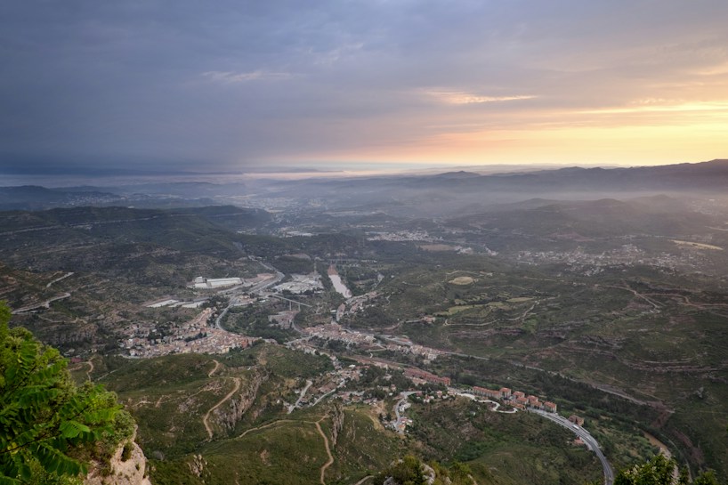 A sprawling landscape viewed from a high vantage point, showcasing a wide valley with a town nestled below. Mountains surround the area, fading into the distance. The sky features a gradient from bright orange and pink near the horizon to dark gray, indicating a sunrise or sunset.