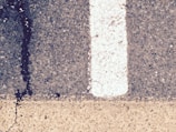 A close-up view of a road surface featuring a white painted line dividing the darker textured asphalt on the right from a lighter-colored strip below. A noticeable crack with a dark stain runs vertically through the surface.