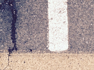 A close-up view of a road surface featuring a white painted line dividing the darker textured asphalt on the right from a lighter-colored strip below. A noticeable crack with a dark stain runs vertically through the surface.