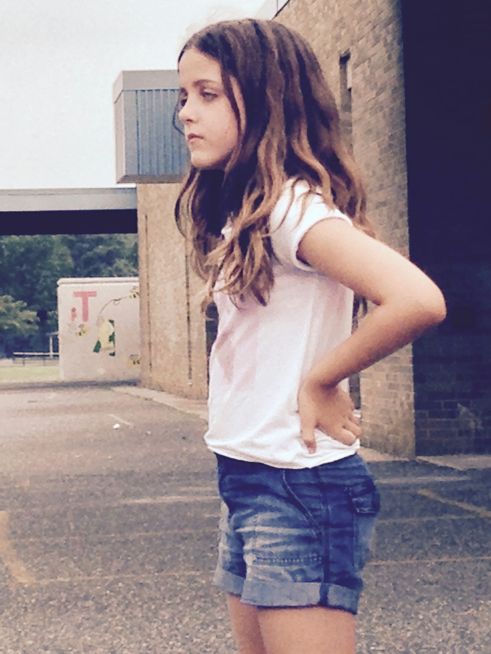 A young girl with long hair standing sideways wearing a white shirt and denim shorts. She has her hands on her hips and appears to be outdoors near a brick building. The background shows a parking area and more buildings.
