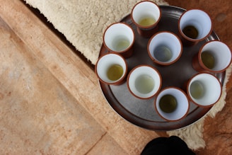 A metal tray holds eight cups with a white interior and brown exterior, each containing a small amount of liquid, possibly tea. The tray rests on a beige textured surface that resembles sheepskin.