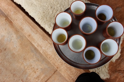 A metal tray holds eight cups with a white interior and brown exterior, each containing a small amount of liquid, possibly tea. The tray rests on a beige textured surface that resembles sheepskin.