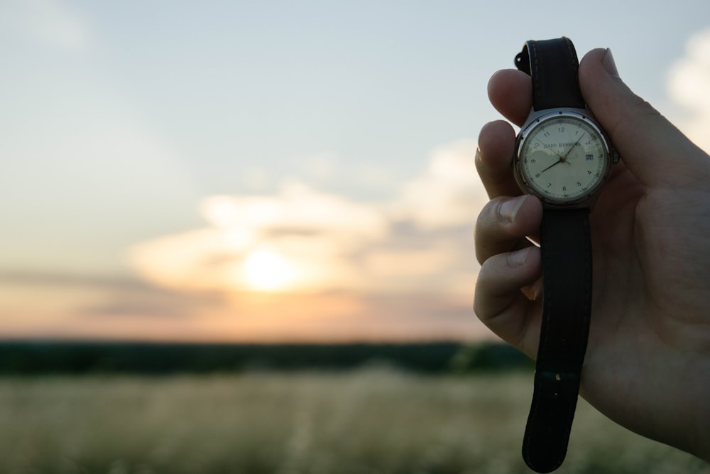 Main d’une personne tenant une montre analogique ronde argentée avec bracelet en cuir noir