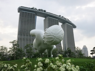 A large white sculpture of a child is positioned on a green landscape with a famous hotel in the background featuring three tower structures connected by a boat-like structure on top. Surrounding the sculpture are vibrant green plants and trees, contributing to a serene and futuristic ambiance.