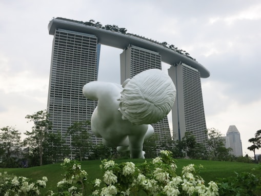 A large white sculpture of a child is positioned on a green landscape with a famous hotel in the background featuring three tower structures connected by a boat-like structure on top. Surrounding the sculpture are vibrant green plants and trees, contributing to a serene and futuristic ambiance.