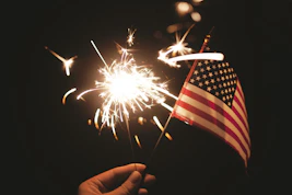 A hand holding a bright sparkler and a small American flag at night