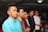 A group of six men stand indoors, dressed in formal attire. They appear to be in a celebratory or formal setting with dim lighting and a decorative background, possibly balloons.