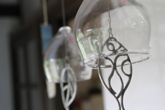 Glass wind chimes hang delicately, capturing a soft and blurred background. Metal crafted figures dangle inside, providing intricate details within each transparent chime.