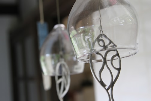 Glass wind chimes hang delicately, capturing a soft and blurred background. Metal crafted figures dangle inside, providing intricate details within each transparent chime.