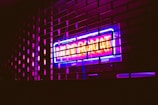 A brightly lit neon sign in vibrant colors shines through a metal grill. The sign reads 'Laundrymat' in eye-catching blue and pink hues, casting reflections on the surrounding area and creating an urban, night-time atmosphere.