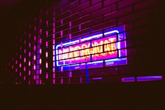 A brightly lit neon sign in vibrant colors shines through a metal grill. The sign reads 'Laundrymat' in eye-catching blue and pink hues, casting reflections on the surrounding area and creating an urban, night-time atmosphere.