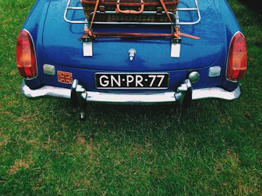 A classic blue car with a rear luggage rack containing a wicker basket. The license plate reads 'GN-PR-77' and a decal with the Union Jack is visible. It is parked on a grassy surface, with moisture indicating recent rain.